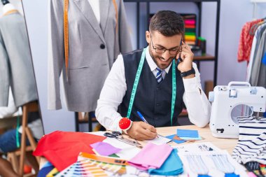 Genç İspanyol terzi terzide akıllı telefondan konuşuyor. Terzide defter üzerinde yazıyor.