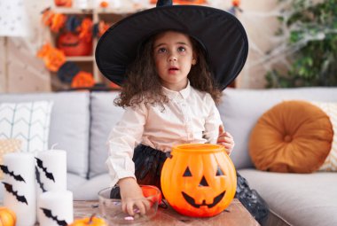 Adorable hispanic girl having halloween party putting sweets on pumpkin basket at home