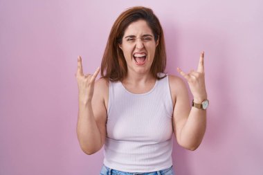 Brunette woman standing over pink background shouting with crazy expression doing rock symbol with hands up. music star. heavy music concept. 