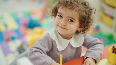 Adorable hispanic girl preschool student sitting on table drawing on paper at kindergarten