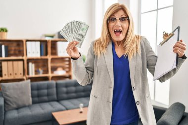 Middle age blonde woman working at therapy consultation office holding money celebrating crazy and amazed for success with open eyes screaming excited. 
