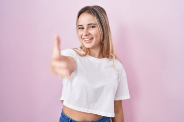 Young blonde woman standing over pink background smiling friendly offering handshake as greeting and welcoming. successful business. 