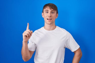 Caucasian blond man standing over blue background pointing finger up with successful idea. exited and happy. number one. 