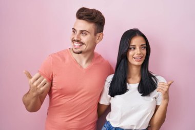 Pembe arka planda duran genç İspanyol çift mutlu yüzle gülümsüyor ve başparmağıyla yan tarafa bakıyor.. 
