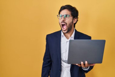 Handsome latin man working using computer laptop angry and mad screaming frustrated and furious, shouting with anger. rage and aggressive concept. 