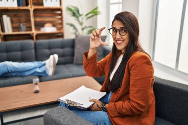 Young hispanic woman working as psychology counselor smiling and confident gesturing with hand doing small size sign with fingers looking and the camera. measure concept. 
