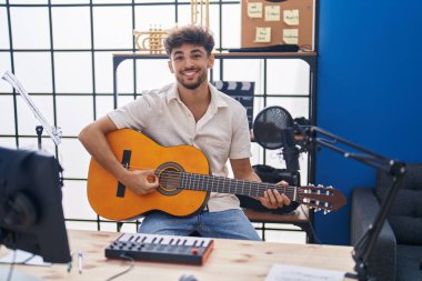 Arab man with beard playing classic guitar at music studio looking positive and happy standing and smiling with a confident smile showing teeth 
