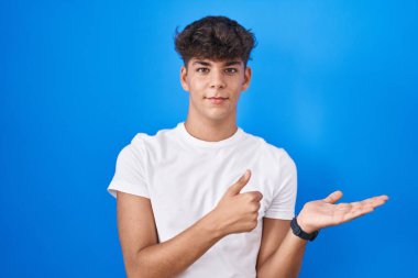 Hispanic teenager standing over blue background showing palm hand and doing ok gesture with thumbs up, smiling happy and cheerful 