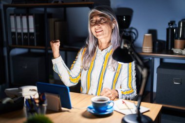 Gri saçlı orta yaşlı bir kadın gece ofiste çalışıyor. Gülümsüyor, gülümsüyor ve baş parmağıyla yüzünü gösteriyor.. 