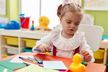 Sevimli sarışın anaokulu öğrencisi anaokulunda masaya oturmuş kağıda resim çiziyor.