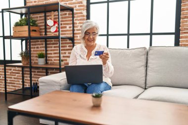 Evdeki koltukta oturan orta yaşlı bir kadın dizüstü bilgisayar ve kredi kartı kullanıyor.