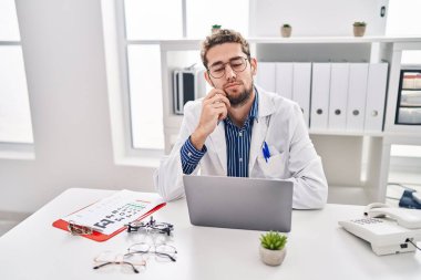 Hispanic man with beard wearing doctor uniform and stethoscope thinking concentrated about doubt with finger on chin and looking up wondering 