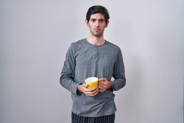 Young hispanic man wearing pajama drinking a cup of coffee skeptic and nervous, frowning upset because of problem. negative person. 