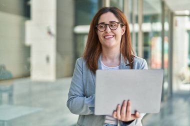 Genç iş kadını sokakta dizüstü bilgisayar kullanarak gülümsüyor.