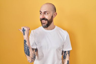 Sarı arka planda dikilen, gülümseyen, gülümseyen ve baş parmağıyla yüzünü gösteren İspanyol dövmeli genç adam.. 