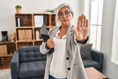 Middle age woman with grey hair at consultation office doing stop sing with palm of the hand. warning expression with negative and serious gesture on the face. 