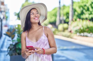 Genç, güzel İspanyol kadın turist parkta akıllı telefon kullanarak gülümsüyor.