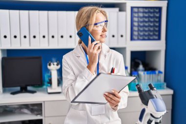 Young blonde woman scientist talking on the smartphone at laboratory