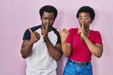 Genç Afro-Amerikan çift pembe arka planda dikiliyorlar ve sessiz olmak istiyorlar. Parmakları dudaklarında, elleri yana doğru işaret ediyorlar. sessizlik ve gizli konsept. 