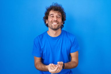 Hispanic young man standing over blue background with hands together and crossed fingers smiling relaxed and cheerful. success and optimistic 