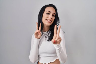Hispanic woman standing over isolated background smiling looking to the camera showing fingers doing victory sign. number two. 