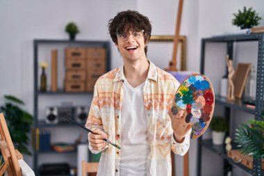 Young hispanic man artist holding paintbrush and palette at art studio