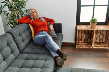 Orta yaşlı, kır saçlı, gülümseyen bir kadın. Evdeki kanepede oturuyor.