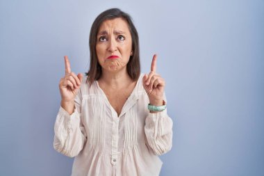 Orta yaşlı İspanyol kadın mavi arka planda dikiliyor üzgün ve üzgün görünüyor, parmaklarıyla yön gösteriyor, mutsuz ve depresif.. 