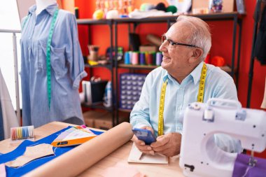 Middle age grey-haired man tailor smiling confident using smartphone at atelier