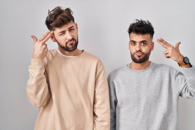 Young homosexual couple standing over white background shooting and killing oneself pointing hand and fingers to head like gun, suicide gesture. 