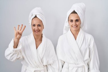 Middle age woman and daughter wearing white bathrobe and towel showing and pointing up with fingers number four while smiling confident and happy. 