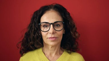 Middle age hispanic woman standing with serious expression over isolated red background