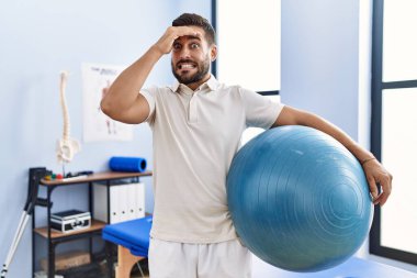 Handsome hispanic man holding pilates ball at rehabilitation clinic stressed and frustrated with hand on head, surprised and angry face 