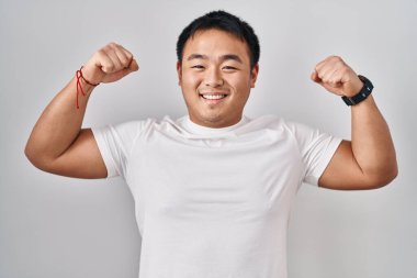 Young chinese man standing over white background showing arms muscles smiling proud. fitness concept. 