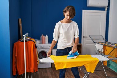 Genç ve güzel İspanyol kadın gülümsüyor çamaşırhanede kendine güvenen ütü yapıyor.