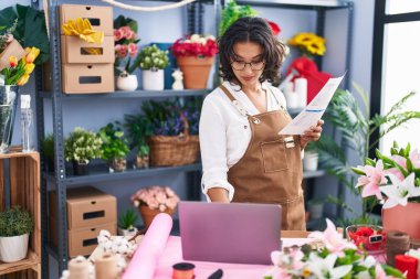 Young beautiful hispanic woman florist using laptop reading document at flower shop