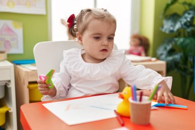 Anaokulunda elinde makasla masada oturan sevimli sarışın çocuk.