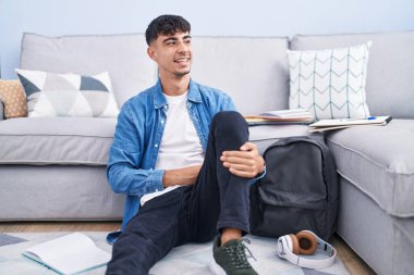 Young hispanic man sitting on the floor studying for university looking away to side with smile on face, natural expression. laughing confident. 