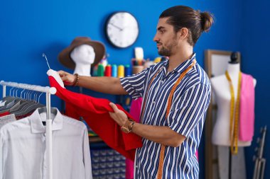 Young hispanic man tailor holding t shirt on rack at sewing studio
