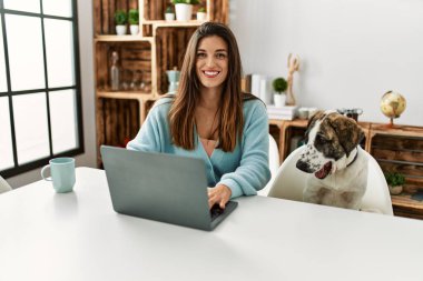 Evde köpekle aynı masada oturan dizüstü bilgisayar kullanan genç bir kadın.
