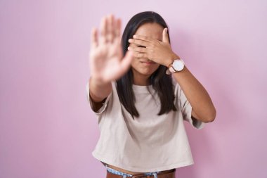 Pembe arka planda duran genç İspanyol kadın elleriyle gözlerini kapatıyor ve üzgün ve korku ifadesiyle dur hareketi yapıyor. utangaç ve olumsuz kavram. 