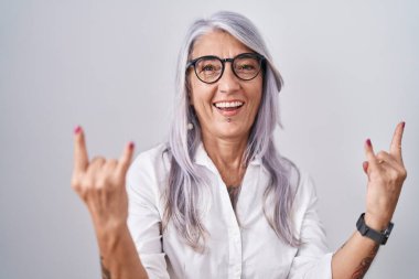 Beyaz arka planda duran gözlüklü orta yaşlı bir kadın çılgın bir ifadeyle el kaldırarak rock sembolü yapıyor. Müzik yıldızı. ağır kavram. 