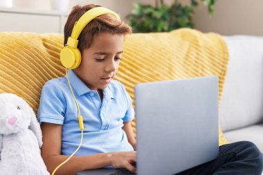 Adorable hispanic boy using laptop and headphones sitting on sofa at home