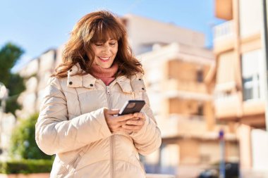 Sokakta akıllı telefon kullanan orta yaşlı bir kadın gülümsüyor.