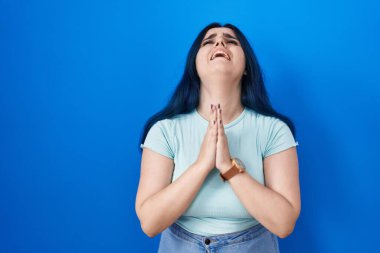 Young modern girl with blue hair standing over blue background begging and praying with hands together with hope expression on face very emotional and worried. begging. 