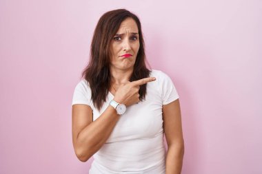 Middle age brunette woman standing over pink background pointing aside worried and nervous with forefinger, concerned and surprised expression 