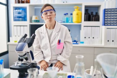 Hispanic girl with down syndrome working at scientist laboratory making fish face with lips, crazy and comical gesture. funny expression. 