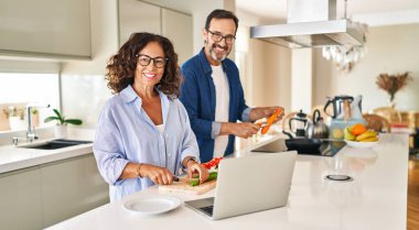 Orta yaşlı İspanyol çift mutfakta yemek pişiriyor ve laptop kullanıyor.