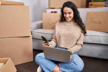 Young hispanic woman using laptop and smartphone sitting on floor at new home