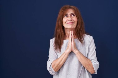 Middle age woman standing over blue background begging and praying with hands together with hope expression on face very emotional and worried. begging. 